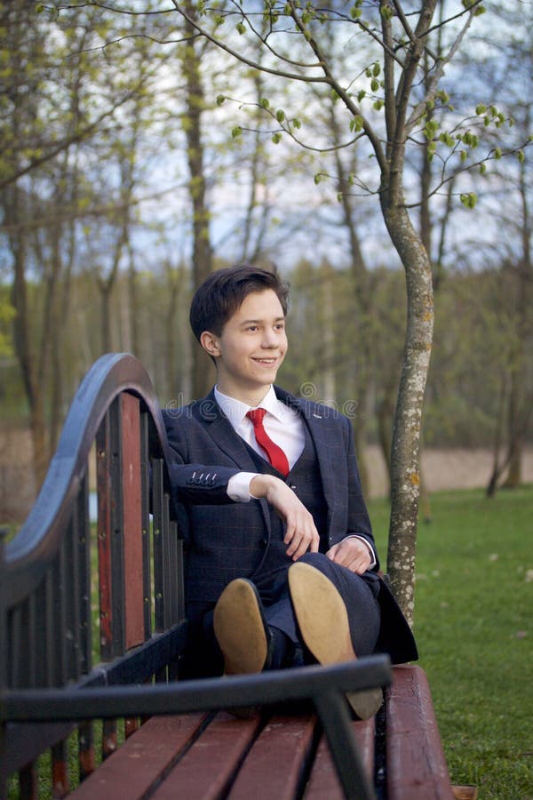Um homem novo, um adolescente, em um terno clássico Senta-se em um banco do vintage em um parque da mola imagens de stock Um homem novo, um adolescente, em um terno clássico Senta-se em um banco do vintage em um parque da mola imagens de stock