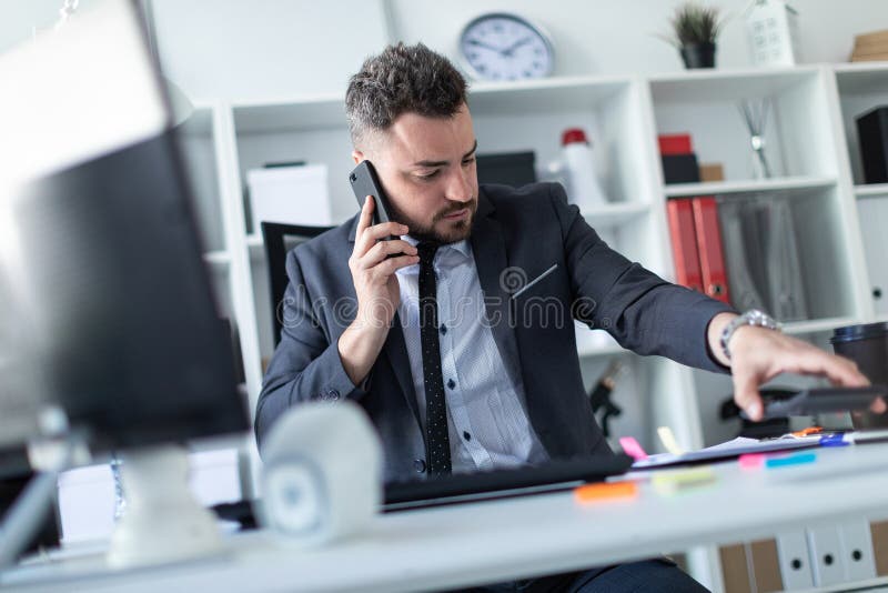 Um homem senta-se no escritório na mesa, fala-se no telefone, trabalha-se com originais e põe-se de lado a calculadora imagem de stock royalty free Um homem senta-se no escritório na mesa, fala-se no telefone, trabalha-se com originais e põe-se de lado a calculadora imagem de stock royalty free
