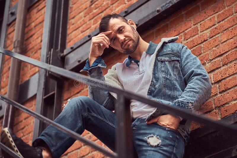 Um homem à moda considerável que veste um revestimento da sarja de Nimes que levanta em escadas fora, olhando uma câmera foto de stock royalty free Um homem à moda considerável que veste um revestimento da sarja de Nimes que levanta em escadas fora, olhando uma câmera foto de stock royalty free