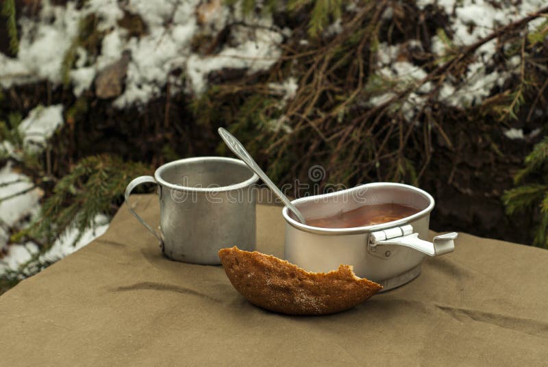 A scanty road meal: soup in a mess tin, a drink in a vintage army mug, and a loaf of bread on a spread tarpaulin on the background of fir branches. A scanty road meal: soup in a mess tin, a drink in a vintage army mug, and a loaf of bread on a spread tarpaulin on the background of fir branches