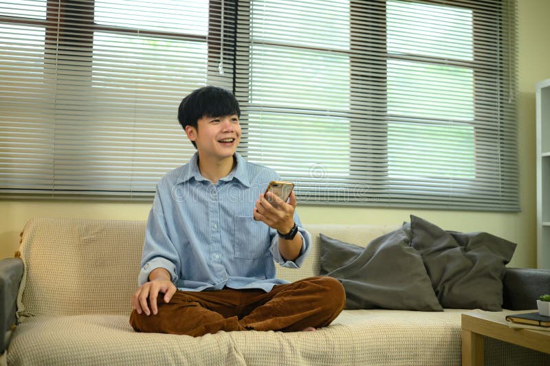 Um jovem sorridente sentado num sofá em casa segurando um smartphone e conversando foto de stock royalty free