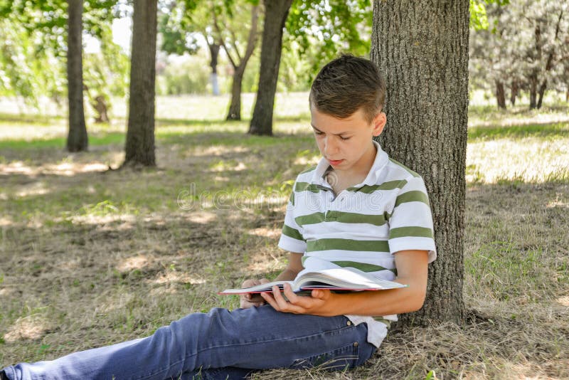 A boy with a textbook under a tree is preparing for school assignments and doing lessons in nature in the park. A boy with a textbook under a tree is preparing for school assignments and doing lessons in nature in the park.