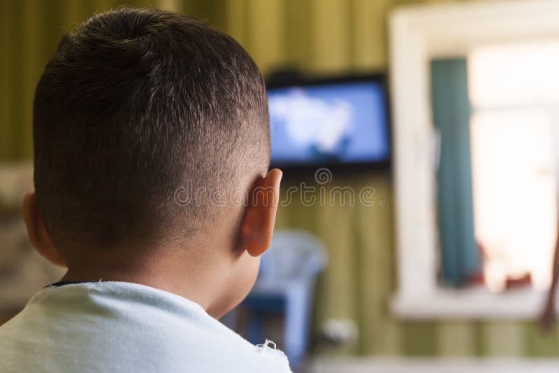 Um menino novo está olhando uma tela da televisão com seu para trás para um efeito da tevê em crianças ou em um conceito de uma c foto de stock Um menino novo está olhando uma tela da televisão com seu para trás para um efeito da tevê em crianças ou em um conceito de uma c foto de stock