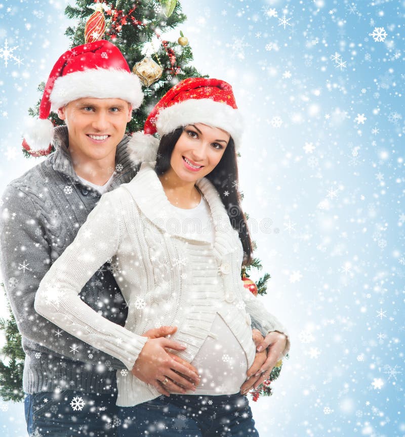 A young and pregnant women and happy father decorating the Christmas tree. The image is taken on a blue background with snow. A young and pregnant women and happy father decorating the Christmas tree. The image is taken on a blue background with snow.