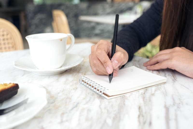 Um ` s da mulher entrega a escrita para baixo em um caderno vazio branco com copo e sobremesa de café na tabela no café imagem de stock royalty free Um ` s da mulher entrega a escrita para baixo em um caderno vazio branco com copo e sobremesa de café na tabela no café imagem de stock royalty free