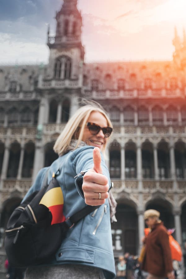 Um viajante fêmea com uma trouxa e uma bandeira de Bélgica está no quadrado de Grand Place em Bruxelas e mostra seus polegares ac imagem de stock Um viajante fêmea com uma trouxa e uma bandeira de Bélgica está no quadrado de Grand Place em Bruxelas e mostra seus polegares ac imagem de stock