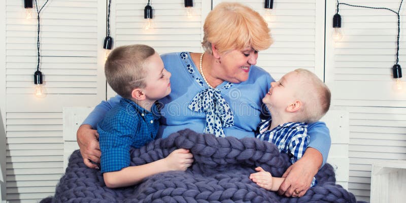 Uma avó bonita e dois netos estão sentando-se no sofá sob uma cobertura merino feita malha de lãs Uma família feliz fotografia de stock Uma avó bonita e dois netos estão sentando-se no sofá sob uma cobertura merino feita malha de lãs Uma família feliz fotografia de stock