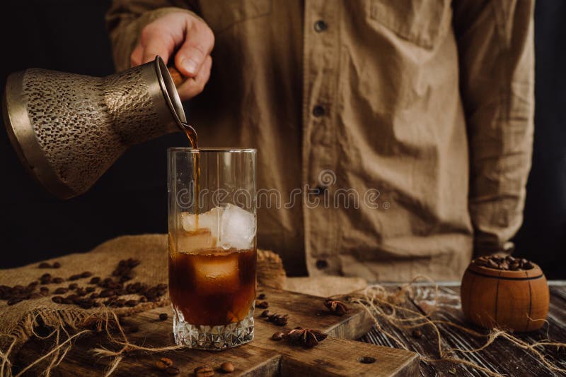 a male barista pours coffee from a metal mug into a glass with ice. a male barista pours coffee from a metal mug into a glass with ice