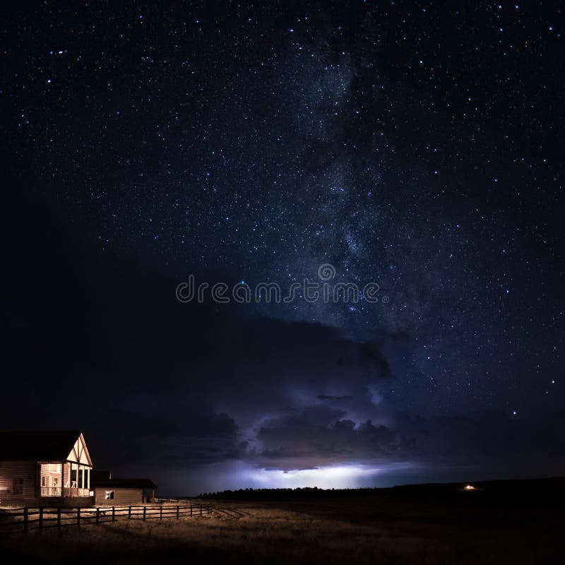 Uma cabine com a luz da varanda acesa e a vedação nos campos com a forma leitosa na noite estrelada acima, juntamente com um raio