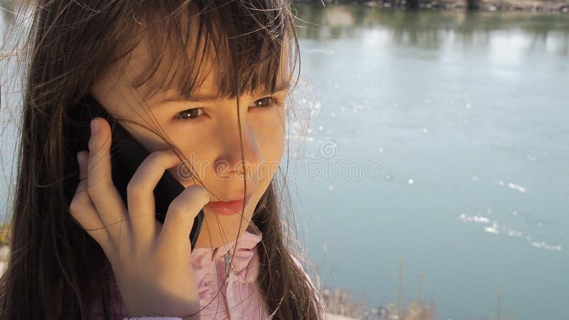 Uma criança com um telefone celular pelo rio A menina está falando no telefone no ar fresco Um dia ensolarado da mola