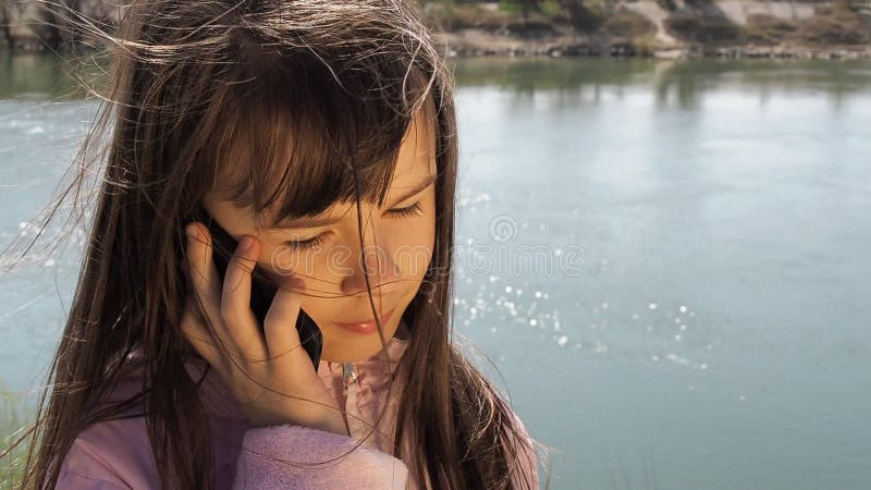 Uma criança com um telefone celular pelo rio A menina está falando no telefone no ar fresco Um dia ensolarado da mola