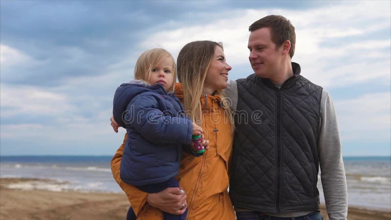 Uma família nova com uma criança pequena que anda ao longo da praia em um dia frio