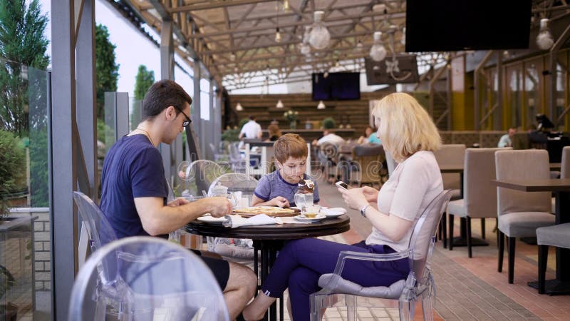 Uma família nova janta em um restaurante, uma mulher é fotografada por seu marido, que bebe o chá, suas bebidas a da criança