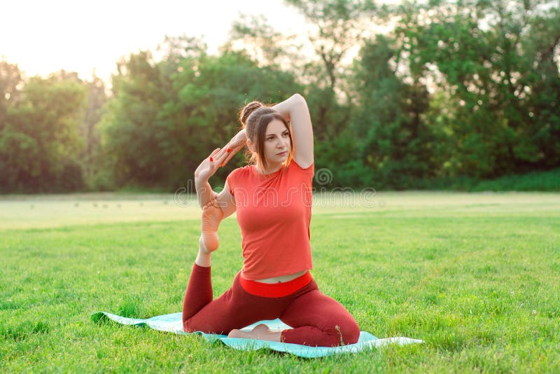 Be fit and healthy. A young girl in orange sportswear on a yoga mat practices yoga in a dove pose with a shin grab. Stretching lessons in the morning at sunrise on the green grass on the soccer field. Be fit and healthy. A young girl in orange sportswear on a yoga mat practices yoga in a dove pose with a shin grab. Stretching lessons in the morning at sunrise on the green grass on the soccer field.