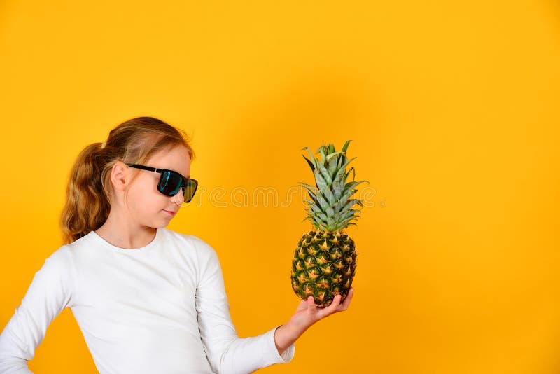 Uma menina bonita e bonito nos óculos de sol guarda um abacaxi e olha-o, em um fundo amarelo no estúdio foto de stock royalty free Uma menina bonita e bonito nos óculos de sol guarda um abacaxi e olha-o, em um fundo amarelo no estúdio foto de stock royalty free