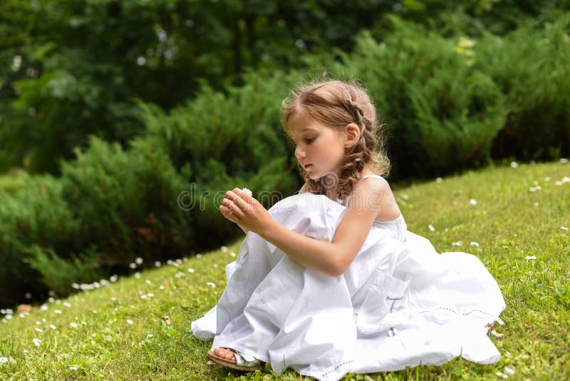 Uma menina bonita em um vestido longo branco que senta-se na grama e na vista verde-clara fotografia de stock royalty free Uma menina bonita em um vestido longo branco que senta-se na grama e na vista verde-clara fotografia de stock royalty free