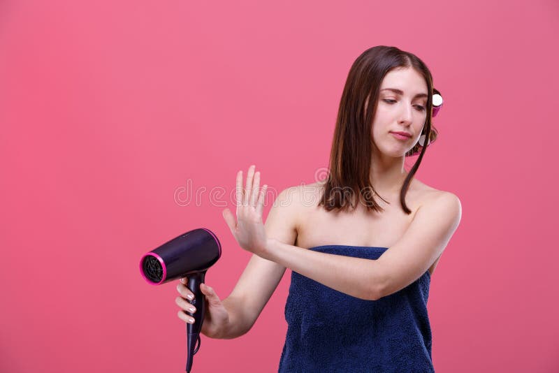 Uma menina, com encrespadores de cabelo e envolvido em uma toalha de banho, guarda um hairdryer e gerencie a cara na aversão dela imagens de stock royalty free Uma menina, com encrespadores de cabelo e envolvido em uma toalha de banho, guarda um hairdryer e gerencie a cara na aversão dela imagens de stock royalty free