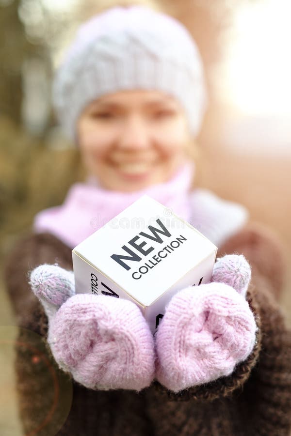 Uma menina de sorriso em um chapéu do inverno e posses dos mitenes em seus braços estendidos um cubo com a coleção nova da inscri imagem de stock Uma menina de sorriso em um chapéu do inverno e posses dos mitenes em seus braços estendidos um cubo com a coleção nova da inscri imagem de stock
