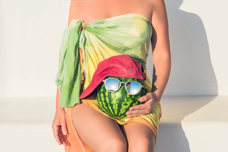 Uma menina despida bonita em um pareo que situa em um banco de pedra branco com uma melancia em um tampão e nos óculos de sol em foto de stock Uma menina despida bonita em um pareo que situa em um banco de pedra branco com uma melancia em um tampão e nos óculos de sol em foto de stock