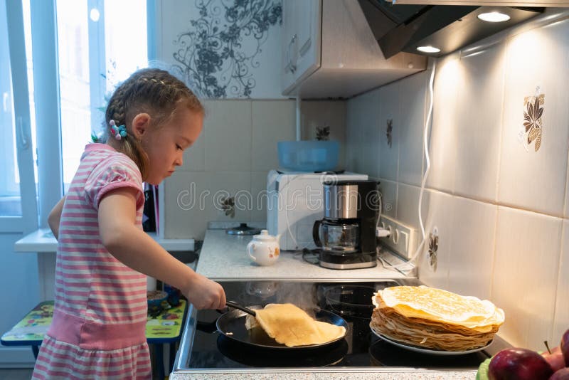 Uma menina em um vestido cor-de-rosa est? fritando panquecas em um fog?o el?trico imagens de stock Uma menina em um vestido cor-de-rosa est? fritando panquecas em um fog?o el?trico imagens de stock
