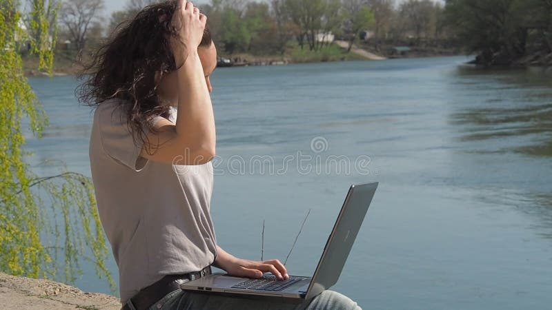 Uma menina está sentando-se com um portátil no banco de rio Um dia ensolarado da mola na margem Jovem mulher na água com um portá