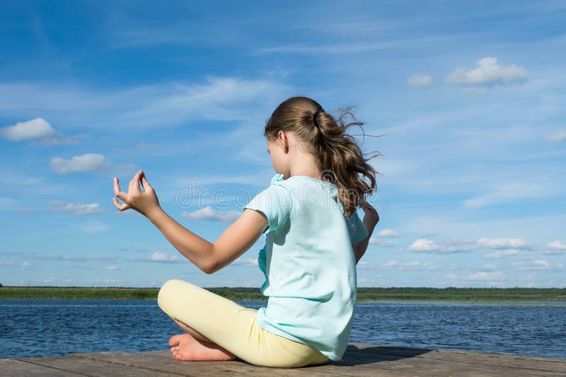 Uma menina está sentando-se na posição dos lótus sobre o cais pelo lago sobre o fundo de um céu azul bonito imagens de stock royalty free Uma menina está sentando-se na posição dos lótus sobre o cais pelo lago sobre o fundo de um céu azul bonito imagens de stock royalty free