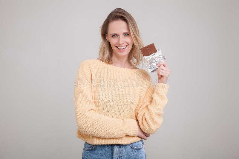 Uma menina macia nova bonita com cabelo louro guarda uma barra de chocolate na cara que est? vestindo em uma camiseta amarela pos foto de stock Uma menina macia nova bonita com cabelo louro guarda uma barra de chocolate na cara que est? vestindo em uma camiseta amarela pos foto de stock