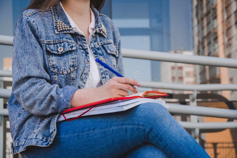 Uma menina na roupa das calças de brim senta-se em um banco, guarda-se cadernos e escreve-se Na rua, escreve em um caderno, estud imagem de stock Uma menina na roupa das calças de brim senta-se em um banco, guarda-se cadernos e escreve-se Na rua, escreve em um caderno, estud imagem de stock