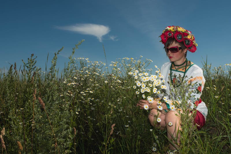 Uma menina no bordado ucraniano com uma grinalda em sua cabeça pelo lago em um prado entre as flores foto de stock Uma menina no bordado ucraniano com uma grinalda em sua cabeça pelo lago em um prado entre as flores foto de stock