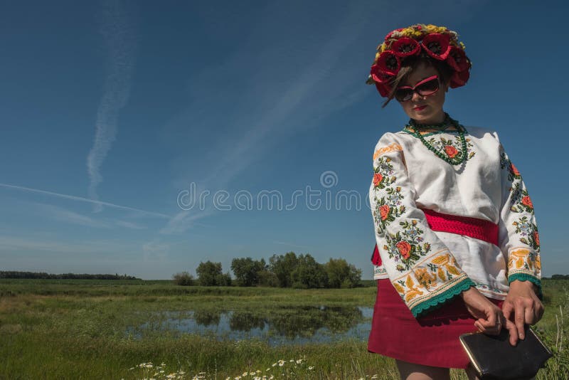 Uma menina no bordado ucraniano com uma grinalda em sua cabeça pelo lago em um prado entre as flores foto de stock Uma menina no bordado ucraniano com uma grinalda em sua cabeça pelo lago em um prado entre as flores foto de stock