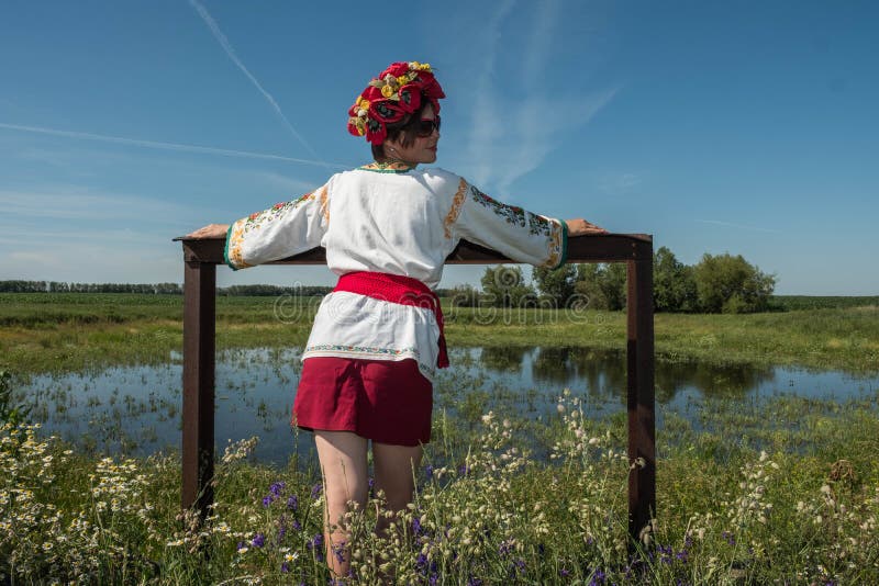 Uma menina no bordado ucraniano com uma grinalda em sua cabeça pelo lago em um prado entre as flores imagens de stock Uma menina no bordado ucraniano com uma grinalda em sua cabeça pelo lago em um prado entre as flores imagens de stock