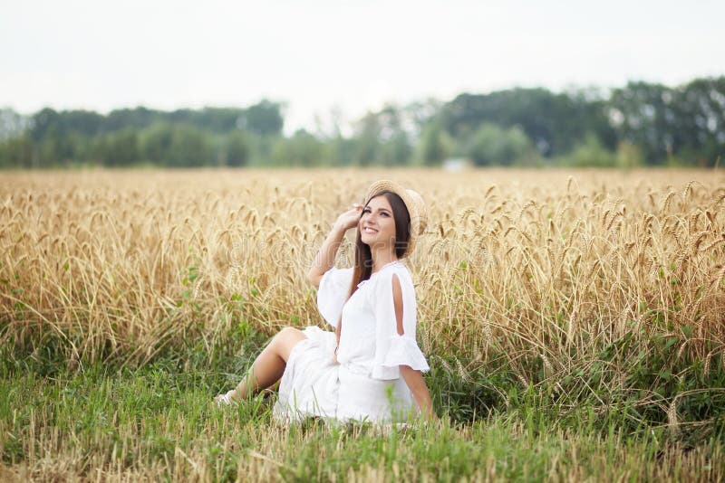 Uma moça em um chapéu é um barqueiro que aprecia a natureza de um campo de trigo Menina bonita nas corridas brancas do vestido no foto de stock Uma moça em um chapéu é um barqueiro que aprecia a natureza de um campo de trigo Menina bonita nas corridas brancas do vestido no foto de stock