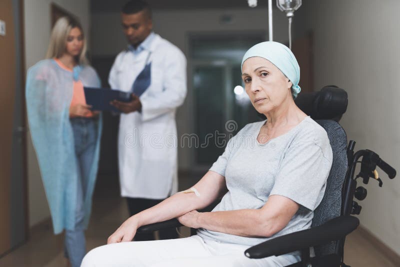 Uma mulher com câncer, com um olhar condenado, está sentando-se em uma cadeira de rodas Atrás de sua filha está e fala ao doutor foto de stock Uma mulher com câncer, com um olhar condenado, está sentando-se em uma cadeira de rodas Atrás de sua filha está e fala ao doutor foto de stock