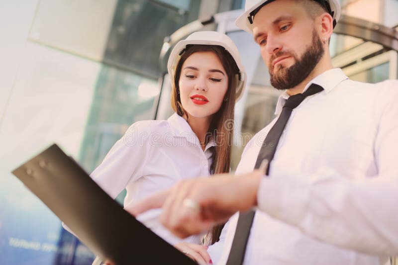 Uma mulher e um homem na roupa do negócio e nos capacetes brancos da construção discutem um plano da construção ou um contrato imagens de stock royalty free Uma mulher e um homem na roupa do negócio e nos capacetes brancos da construção discutem um plano da construção ou um contrato imagens de stock royalty free