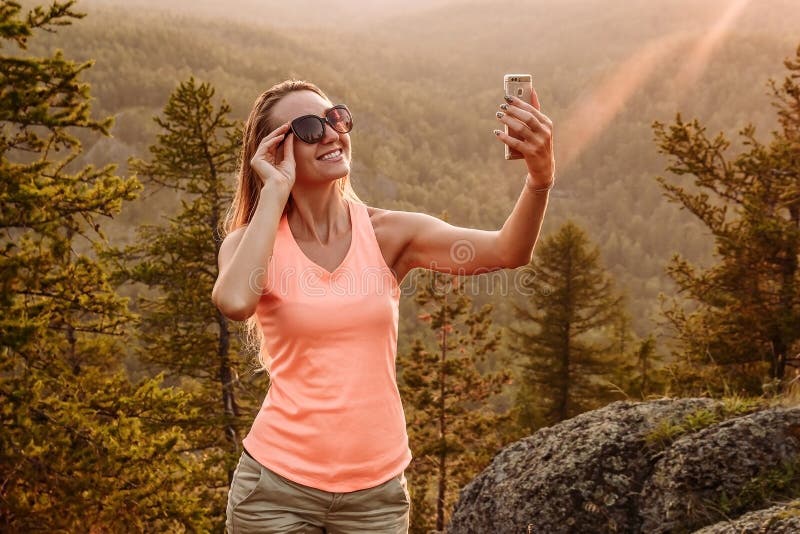 Uma mulher feliz em um t-shirt e no short faz uma foto do selfie nas montanhas no verão Os raios do sol do por do sol imagens de stock royalty free Uma mulher feliz em um t-shirt e no short faz uma foto do selfie nas montanhas no verão Os raios do sol do por do sol imagens de stock royalty free