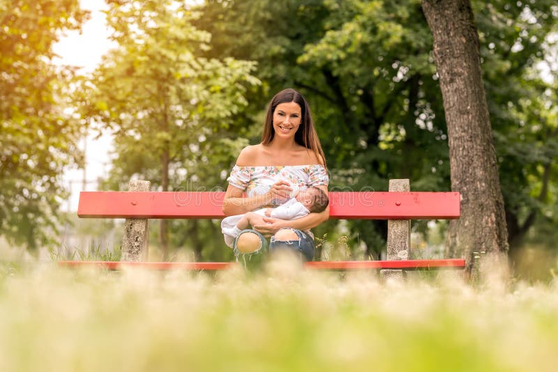 Uma mãe nova que senta-se em um banco e que alimenta seu bebê fotografia de stock Uma mãe nova que senta-se em um banco e que alimenta seu bebê fotografia de stock