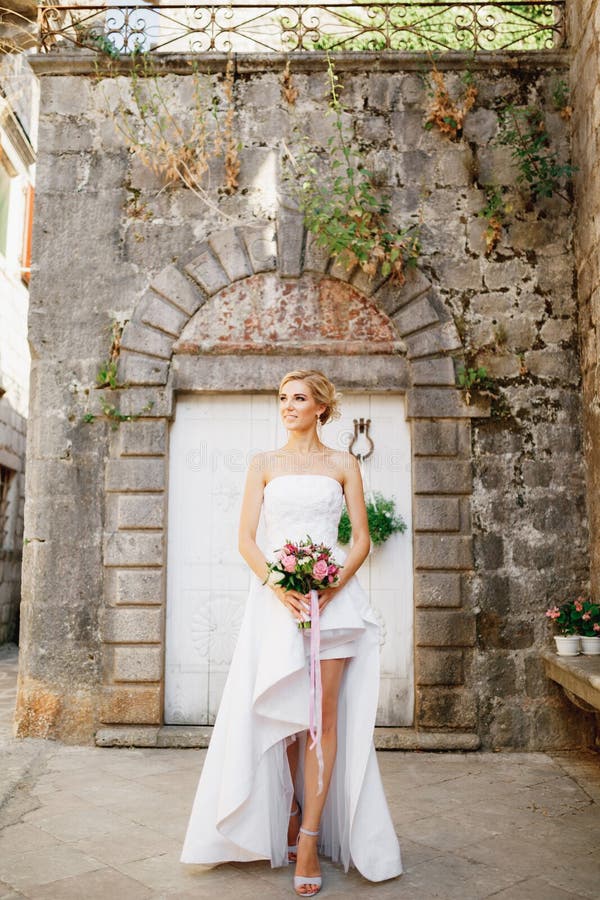 Uma Noiva Com Um Vestido Elegante Com Um Buquê Nas Mãos Está De Pé Nas  Portas Brancas De Um Belo Edifício Antigo Em Perast Imagem de Stock -  Imagem de vestido, senhora: