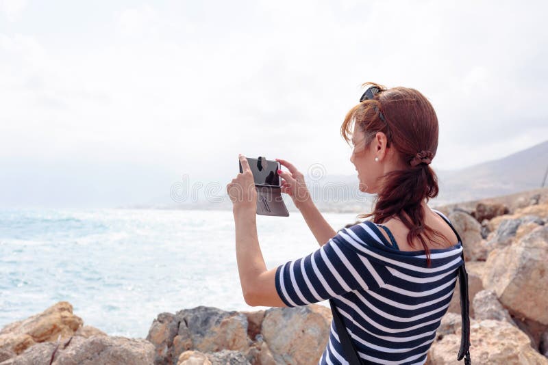 Uma viajante madura atrativa usa um telefone celular para tirar uma foto de uma bela paisagem marinha imagens de stock Uma viajante madura atrativa usa um telefone celular para tirar uma foto de uma bela paisagem marinha imagens de stock