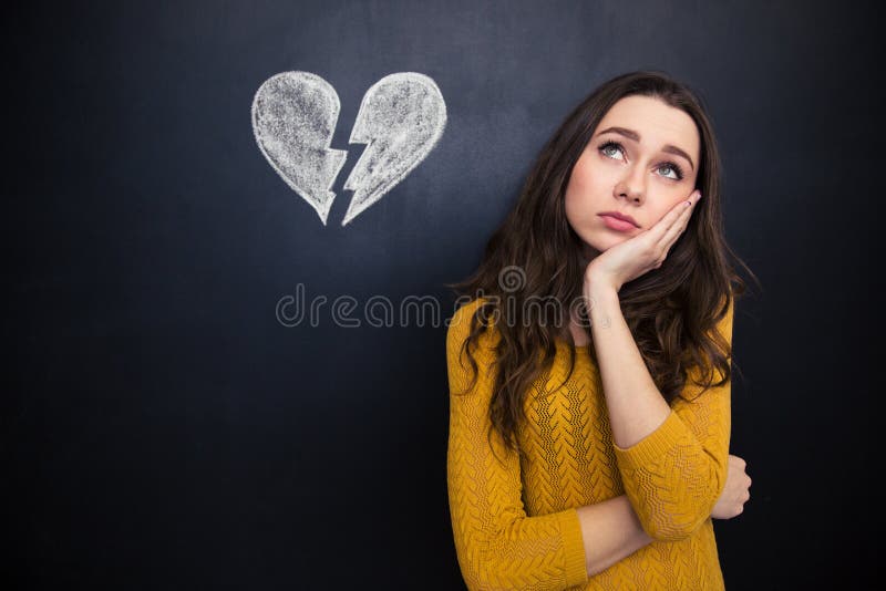 Upset beautiful young woman standing and thinking over background of chalkboard with drawn broken heart. Upset beautiful young woman standing and thinking over background of chalkboard with drawn broken heart