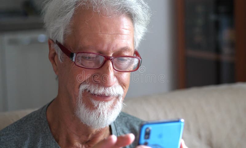 Un anciano de 70 años usando teléfonos inteligentes en línea mirando la pantalla relajándose en casa fotografía de archivo