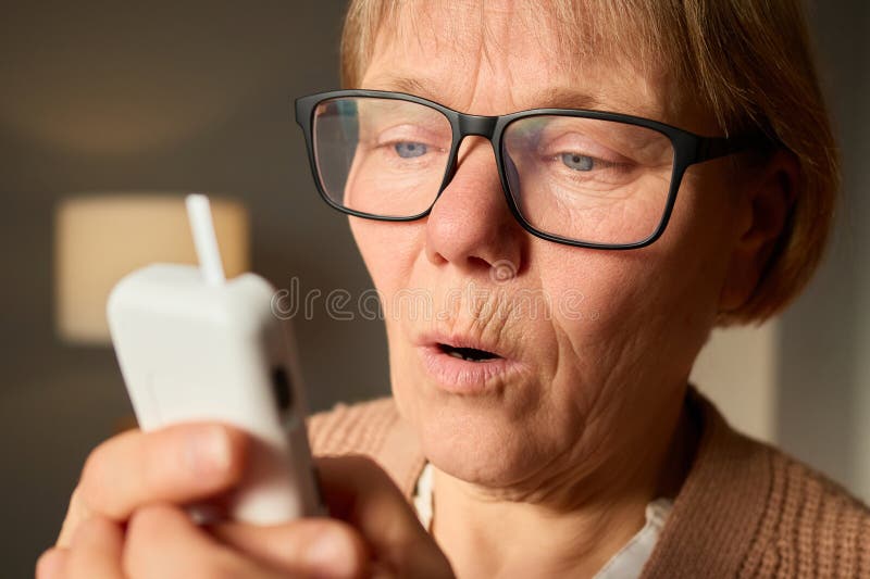 Un'anziana donna sembra stupita mentre tiene il suo smartphone vicino alla sua faccia. La luce calda riempie la camera accogliente immagine stock libera da diritti