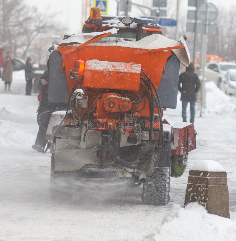 Un arado de nieve está conduciendo por una calle nevada. el arado es de color naranja y tiene un gran cubo de nieve en la parte delantera. hay varias personas caminando por la acera algunos de ellos llevando paraguas