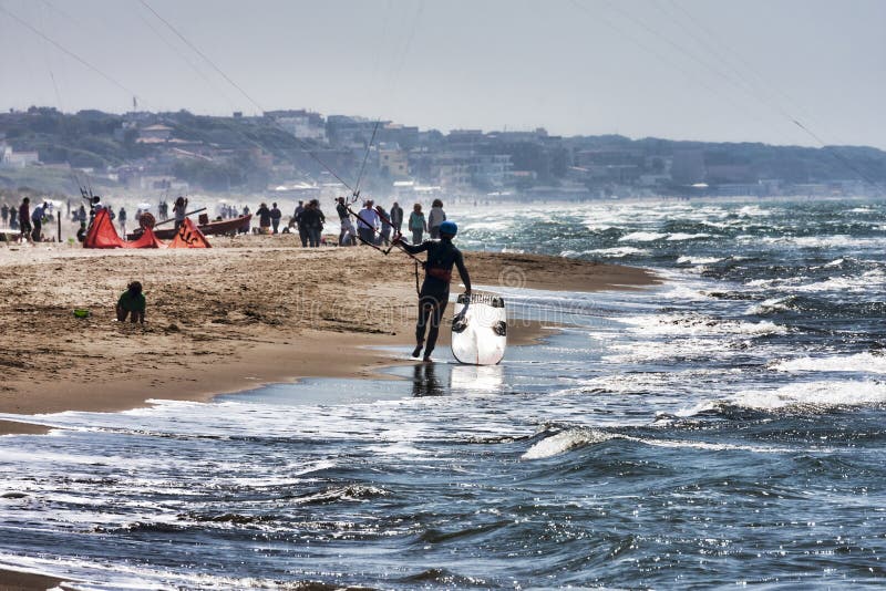 Un atleta camina entre la espuma de las ondas apuntala con su tablero que practica surf de la cometa en la playa romana apretada fotografía de archivo libre de regalías Un atleta camina entre la espuma de las ondas apuntala con su tablero que practica surf de la cometa en la playa romana apretada fotografía de archivo libre de regalías