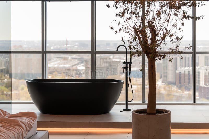 Un bagno spazioso con una vasca da bagno nera elegante accanto a una grande finestra con vista panoramica sulla città. immagini stock