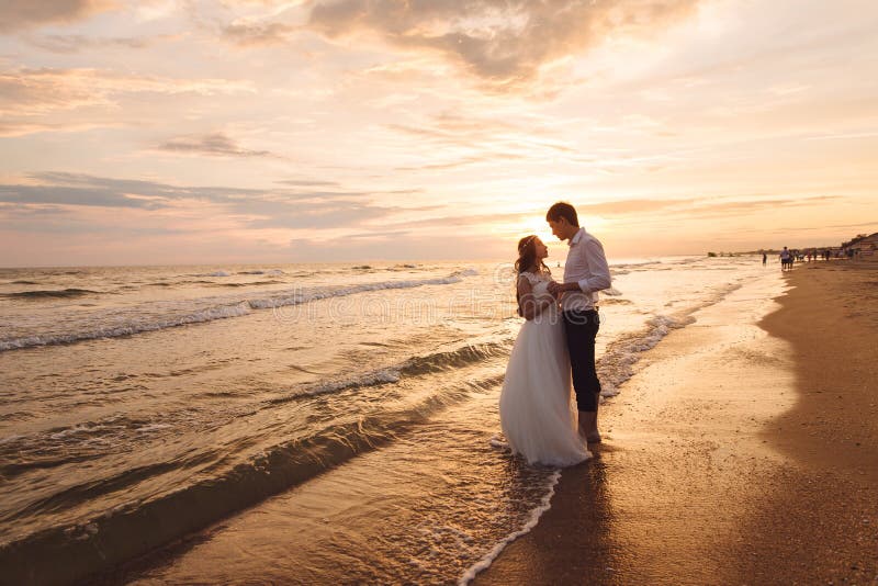 Un beau couple des nouveaux mariés, les jeunes mariés marchant sur la plage Coucher du soleil magnifique et ciel Robes de mariage photographie stock libre de droits Un beau couple des nouveaux mariés, les jeunes mariés marchant sur la plage Coucher du soleil magnifique et ciel Robes de mariage photographie stock libre de droits