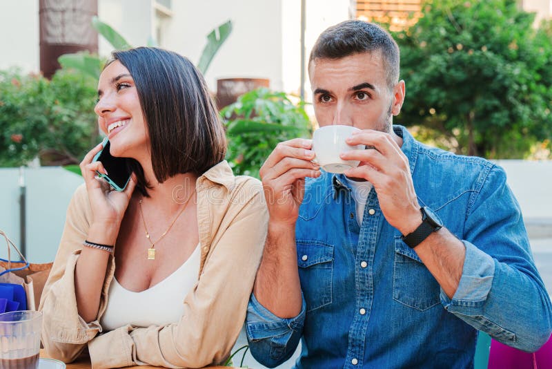 A handsome man drinks and enjoys his coffee cup on a restaurant terrace while his girlsfriend have a phone call conversation. Young adult caucasian couple resting together on a coffeeshop. High quality photo. A handsome man drinks and enjoys his coffee cup on a restaurant terrace while his girlsfriend have a phone call conversation. Young adult caucasian couple resting together on a coffeeshop. High quality photo