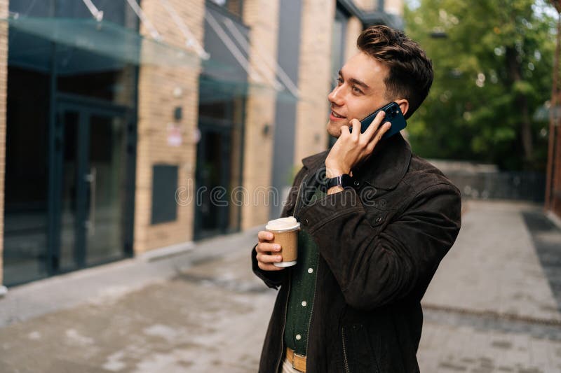 Un beau jeune barbu qui boit un bon café et qui parle sur son smartphone tout en marchant dans la rue de la ville par la ville mod photos stock