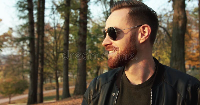 Un beau macho barbu porte des lunettes de soleil regarde en profil et sourit dans le parc