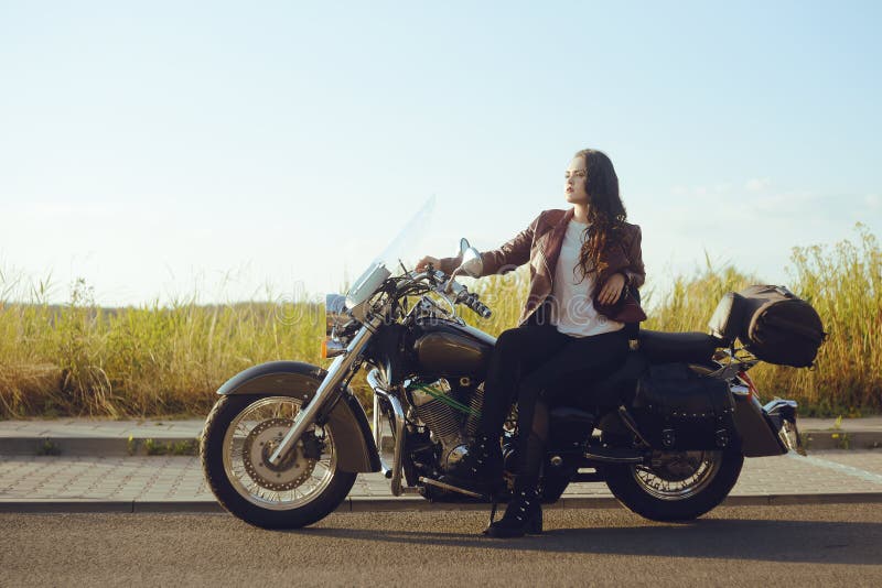 Un bello castana in un bomber rosso sta stando vicino ad un motociclo nel campo Ragazza su un motociclo La ragazza al tramonto immagine stock libera da diritti Un bello castana in un bomber rosso sta stando vicino ad un motociclo nel campo Ragazza su un motociclo La ragazza al tramonto immagine stock libera da diritti