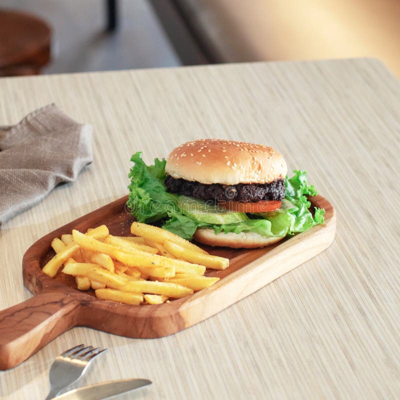 Un burger cu apă dulce, plin cu legume proaspete, stă lângă cartofi prăjiți pe un platou rustic din lemn, într-o atmosferă caldă royalty free stock photos